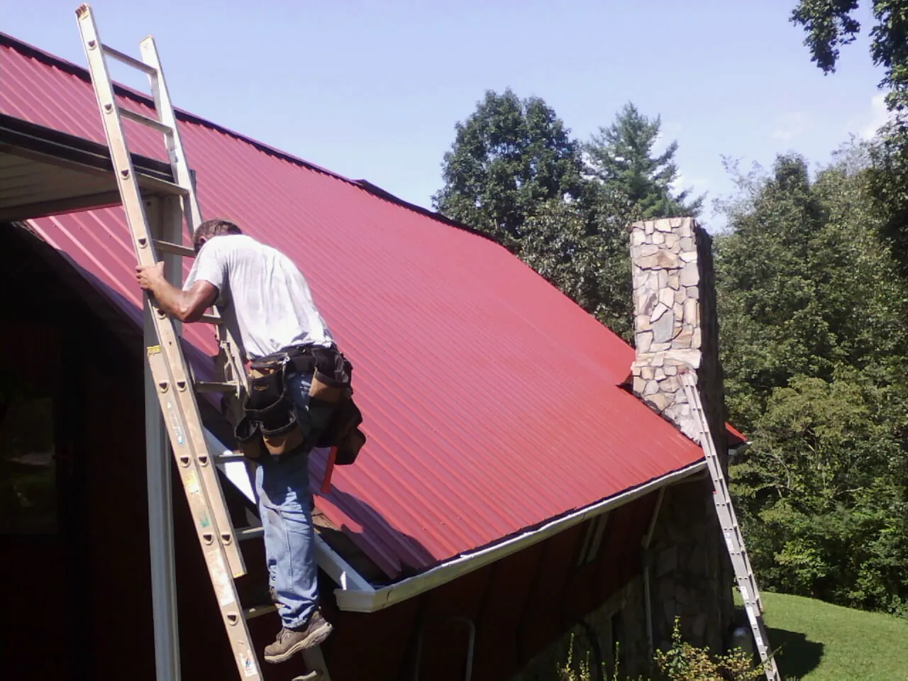 Roofer on a ladder beside a red metal roof and stone chimney — Soffit & Fascia Repair by Roofing Brothers in Mills River